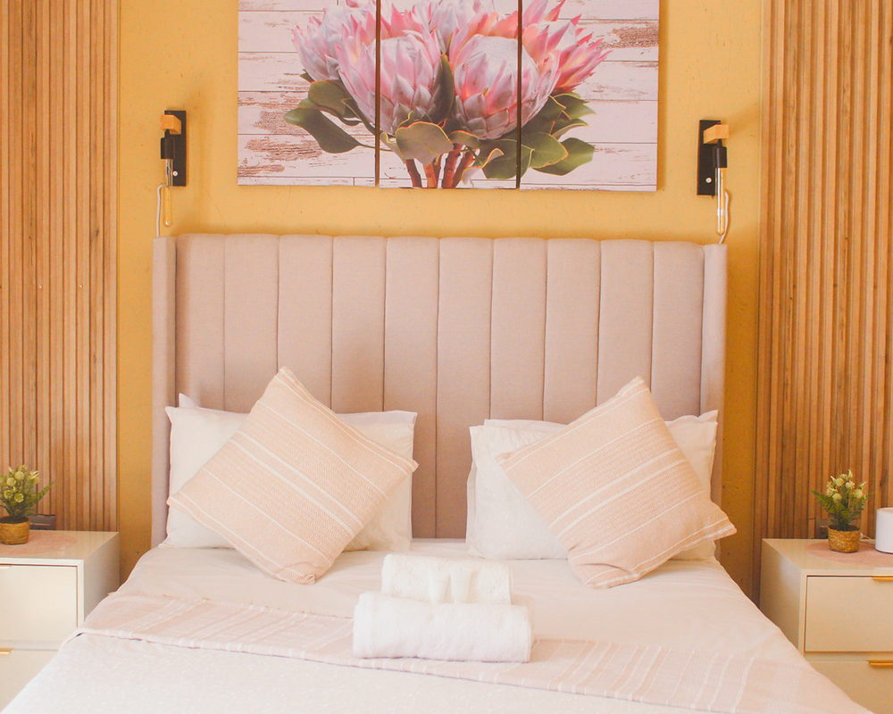 A LAMEMA guest room with an ochre wall, timber panelling and protea artwork above a made-up bed.