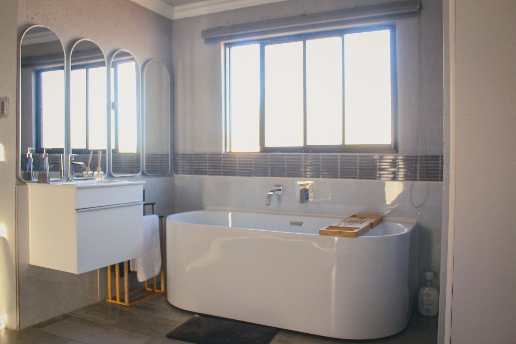 A LAMEMA bathroom with a freestanding oval bath, timber bath caddy and three arched mirrors.