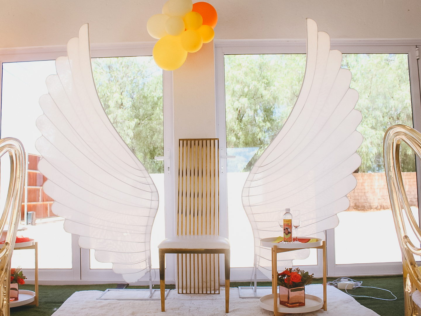 An event styled with a white angel-wing backdrop, an orange and cream balloon garland and gold chairs.