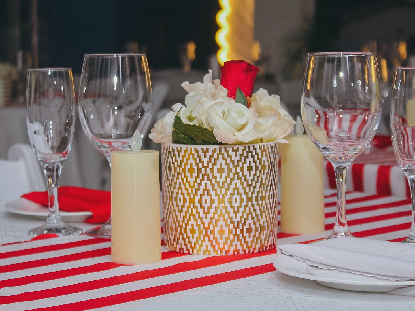 An evening table setting with a red and white striped runner, pillar candles and a red rose in a gold vase.
