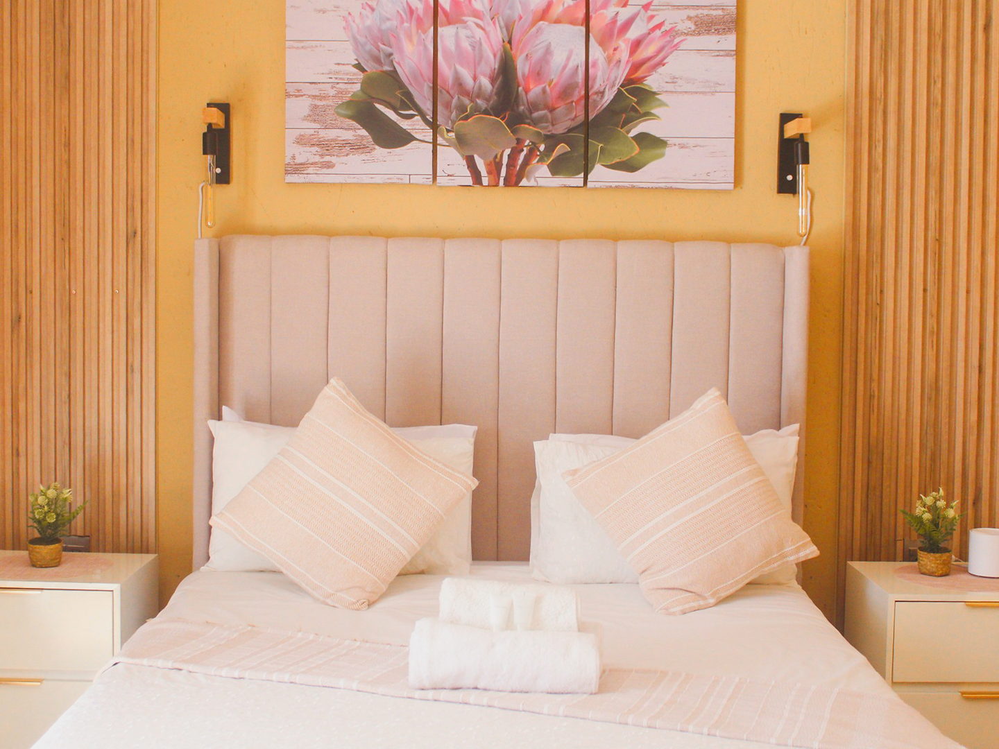 A LAMEMA guest room with an ochre wall, timber panelling and protea artwork above a made-up bed.