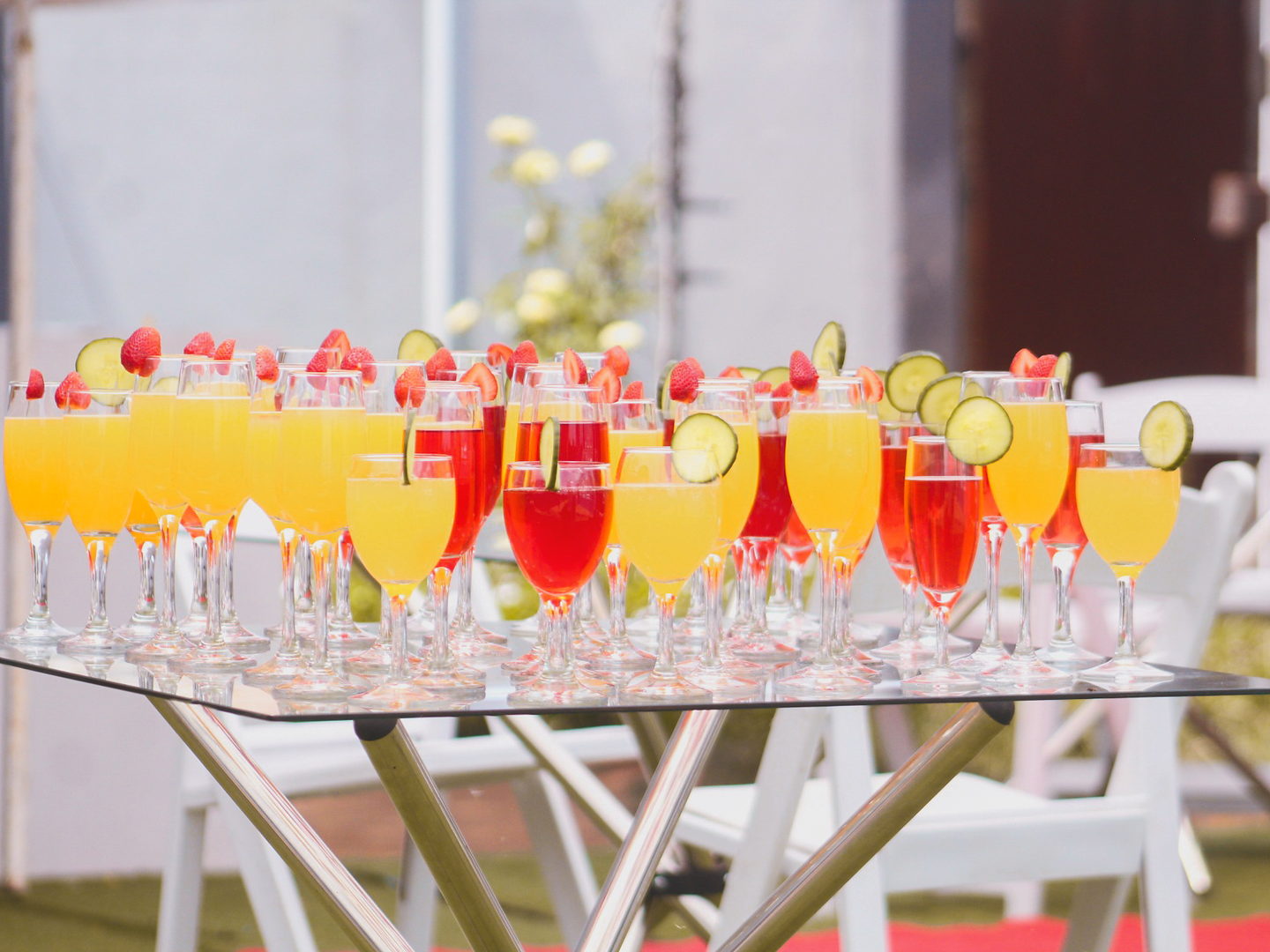 Rows of orange and red welcome cocktails garnished with strawberries and cucumber on a glass table.